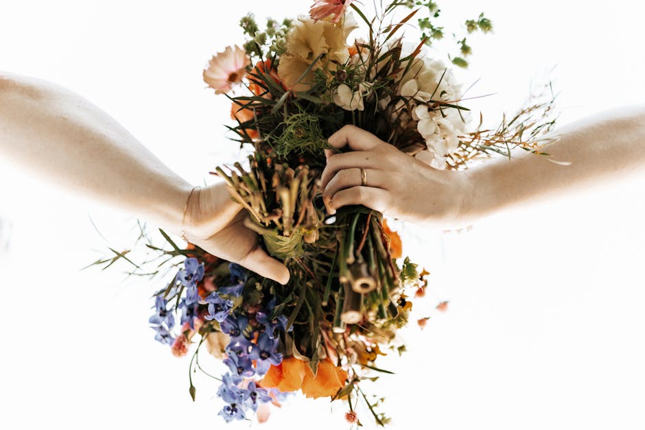 A close-up image of two hands holding a colorful bouquet of fresh flowers, featuring a mix of orange, white, pink, and purple blooms, with green foliage and tendrils. The bouquet is wrapped loosely wi