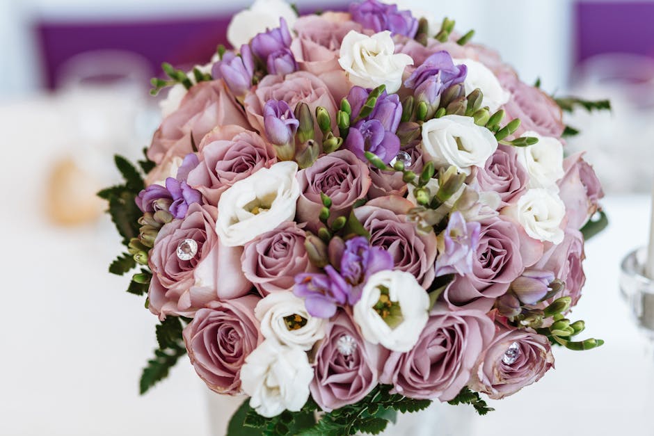 A close-up view of a floral arrangement featuring a round bouquet of fresh, pastel-colored roses in shades of soft pink and lavender, complemented by white lisianthus and purple freesias. The bouquet 