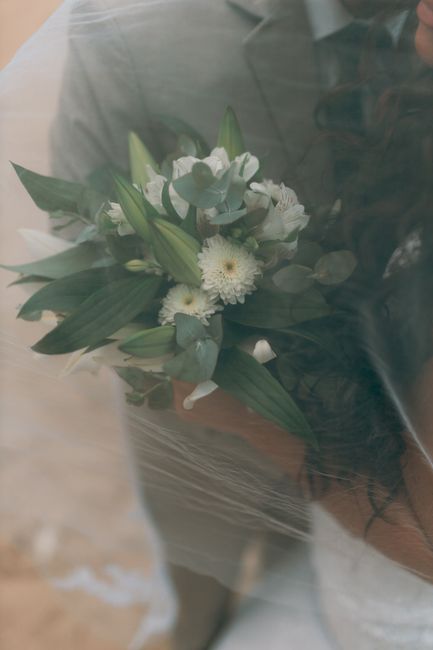 A bouquet of fresh white flowers, including white daisies, lisianthus, and small filler blooms, is arranged with lush green foliage featuring broad and pointed leaves. The bouquet is wrapped in clear 