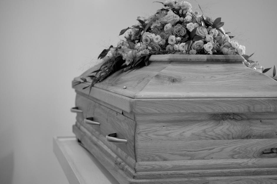 A wooden funeral casket with visible natural grain and smooth polished surface, adorned with a floral arrangement of roses and other flowers in soft pastel shades, resting on a white stand in a softly