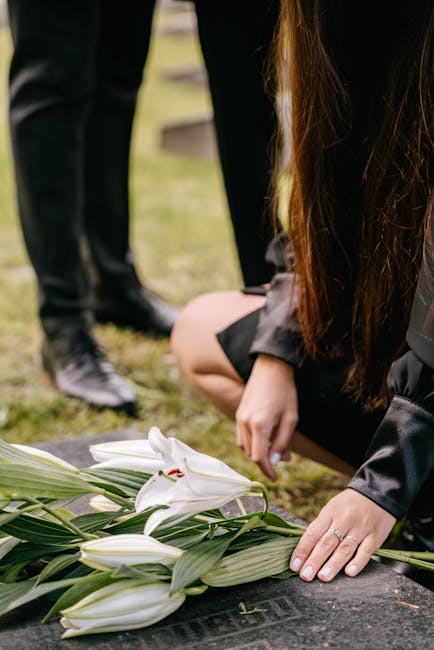 A person dressed in dark clothing is placing a floral arrangement composed of white lilies with large, open petals and glossy green leaves on a memorial stone at a cemetery. The bouquet is wrapped in 