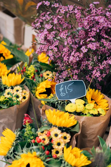 A vibrant display of fresh floral bouquets arranged at a flower market stall, featuring bright yellow sunflowers and small white and yellow daisies wrapped in brown paper. Behind the bouquets, a large
