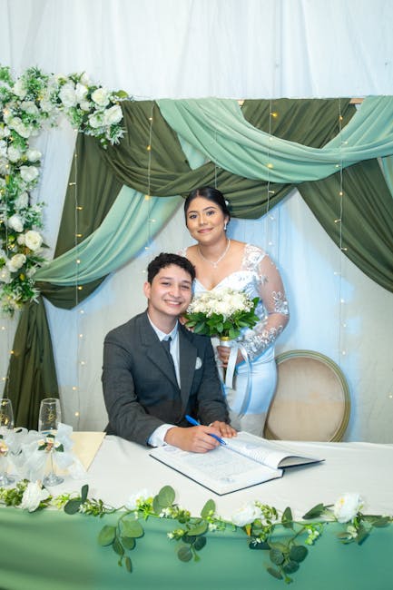 A young woman in a white lace bridal gown holds a bouquet of fresh white roses and greenery, standing behind a smiling young man in a dark suit at a table. The setting features soft pastel green and w